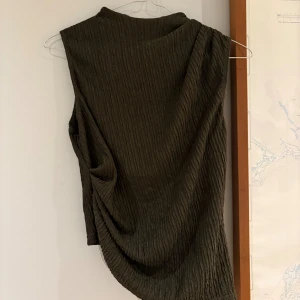 Mörkgrön ärmlös omlotttopp - Säljer en mörkgrön ärmlös topp med omlott-detalj och snyggt strukturerat tyg. Toppen har hög hals och dragkedja bak, samt en asymmetrisk nederkant som ger en cool look. Perfekt för dig som gillar stilrena och unika plagg.