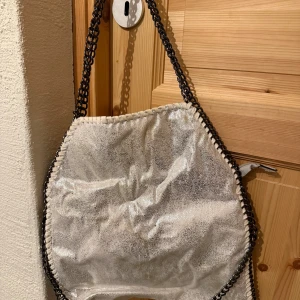 Silver axelväska med kedjedetaljer - Cool axelväska i glittrigt silverfärgat syntetmaterial med grova kedjor som axelrem och längs kanterna. Väskan har dragkedja på baksidan och röd insida. Perfekt för dig som vill sticka ut med en unik accessoar.