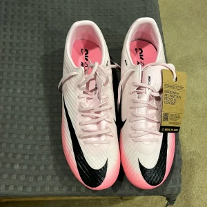 Nike Mercurial rosa fotbollsskor - Nike Mercurial fotbollsskor i ljusrosa med svarta detaljer och stor Swoosh på sidan. Skorna har snörning, rund tå och är tillverkade med minst 20% återvunnet material. Insidan är rosa och yttermaterialet är syntet. Perfekt för dig som vill sticka ut på planen.