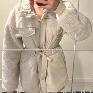 Beige teddyjacka med bälte - Säljer en beige teddyjacka med fluffiga ärmar och axlar, två stora bröstfickor och ett snyggt bälte i midjan. Jackan har hög krage och långa ärmar med ribbade muddar. Perfekt för dig som vill ha en mysig och trendig look.