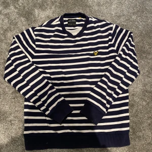 Randig tröja från Lyle & Scott marinblå - Säljer en marinblå och vit randig tröja från Lyle & Scott i storlek M. Tröjan har klassiska horisontella ränder, rund halsringning och den ikoniska gula loggan broderad på bröstet. Tillverkad i 100% bomull och har långa ärmar med ribbade muddar. Har ett litet märke på ryggen men inget man tänker på