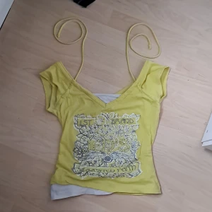 Gul linnetopp från Jennyfer med tryck - Säljer en gul linnetopp från Jennyfer i storlek S. Toppen har smala axelband, v-ringad hals och ett stort blommigt tryck med texten '1st Brand' framtill. Materialet är mjuk bomull och toppen har en figurnära passform. Perfekt för dig som gillar färg och coola detaljer!
