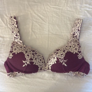 Intimissmi bh  - Säljer en vinröd bh  med ljusa spetsdetaljer i blommigt mönster. Bh  har vadderade kupor och justerbara axelband, samt hak- och hyskknäppning bak. Materialet är mjukt och glansigt med dekorativ spets längs kanterna för extra stil.