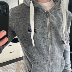 Grå zip hoodie från Superdry - Säljer en grå zip hoodie från Superdry i storlek M, men sitter som en S. Modellen är ca 180cm lång. Hör av dig vid funderingar!