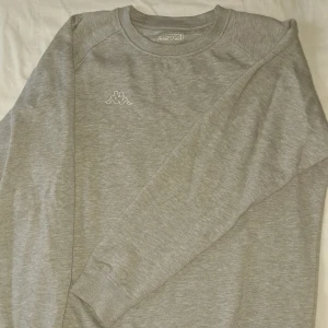 Grå sweatshirt från Kappa - Snygg grå sweatshirt från Kappa med klassisk logga broderad på bröstet. Tröjan har rund hals och långa ärmar, perfekt för chill dagar. Mjuk bomullsmix som känns skön mot huden och passar till jeans eller joggers.