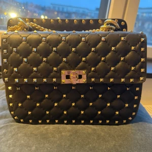 Svart quiltad axelväska från Valentino - Snygg svart axelväska från Valentino med quiltad design och guldiga nitar. Väskan har kedjerem i guld och ett elegant lås framtill. Tillverkad i skinn och perfekt för dig som vill sticka ut med en lyxig accessoar.