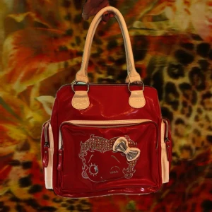 Röd Betty Boop handväska med rosett - Cool röd handväska från Betty Boop med vita detaljer och handtag. Väskan har ett stort fack, flera dragkedjefickor och ett glansigt syntetmaterial. Framsidan pryds av Betty Boop-tryck med glittrande stenar och en 3D-rosett. Perfekt för dig som gillar statement-accessoarer. Lite sönder på ena kanten se bild. 