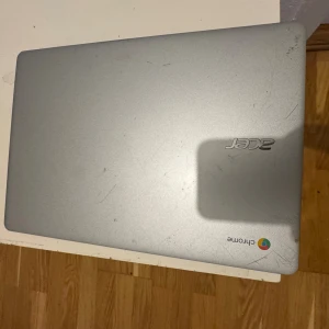 Acer Chromebook - Acer Chromebook med silverfärgat chassi. Synligt slitage och repor på både ovansida och undersida, samt smuts på tangentbordet. Skärmen visar batterisymbol, vilket indikerar att laddning behövs. Perfekt för studier eller surf, säljs i befintligt skick.