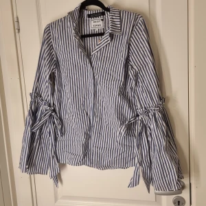 Randig skjorta med knyt från Daisy Grace - Snygg skjorta från Daisy Grace med blåvita ränder och unika långa ärmar med knytband vid armbågen. Skjortan har klassisk krage och är tillverkad i mjuk bomull. Perfekt för dig som gillar detaljer och vill sticka ut lite extra.