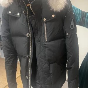 Svart dunjacka Moose Knuckles med päls - Säljer en svart dunjacka från Moose Knuckles med fluffig vit päls på huvan. Jackan har silverfärgade dragkedjor, stora knappar och en snygg logga på ärmen. Flera fickor och quiltade detaljer gör den både praktisk och stilren. Perfekt för kalla dagar.