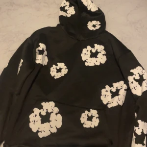 Svart hoodie från Denim Tears med tryck - Svart hoodie från Denim Tears med stora vita blommönster tryckta över hela tröjan. Tröjan har huva och magficka, perfekt för dig som gillar streetwear och vill sticka ut med en unik design.