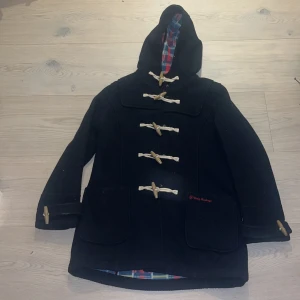 Marinblå duffeljacka från Uniq Outwear - Marinblå duffeljacka med huva från Uniq Outwear, fodrad med färgglatt rutigt tyg i rött, blått och gult. Jackan har klassiska träknappar med snörning, två stora fickor framtill och en loose passform. Perfekt för kalla dagar med sin tjocka ullkänsla.