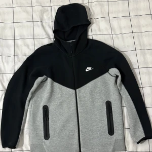 Nike svart och grå hoodiejacka L - Nike hoodiejacka i svart och grått med dragkedja framtill och två fickor med dragkedja. Jackan har huva och är långärmad, med Nike-logga på bröstet. Materialet är en mix av bomull och polyester, vilket gör den mjuk och bekväm. Perfekt för chill och streetwear.