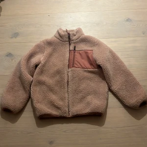 Rosa barnteddyjacka med dragkedja - Mysig barnteddyjacka i rosa med hög krage och hel dragkedja framtill. Jackan har en snygg bröstficka i matchande ton med dragkedja och är helfodrad i samma färg. Perfekt för kalla dagar och har en chill, loose passform.