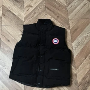 Svart dunväst från Canada Goose - Svart dunväst från Canada Goose med hög krage och klassisk logga på bröstet. Västen har stora fickor framtill och stängs med både dragkedja och knappar. Perfekt för lager-på-lager och håller dig varm under kyliga dagar.