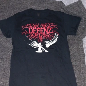 Svart DEFENZ t-shirt med tryck - Svart t-shirt från Gildan med stort rött och vitt tryck på bröstet. Trycket har texten DEFENZ i röd, grenliknande stil och en vit figur med vingar under. Klassisk passform och rund hals.