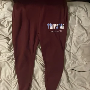 Vinröda Trapstar mjukisbyxor L - Säljer ett par vinröda mjukisbyxor från Trapstar med tryck på ena benet. Byxorna är tillverkade i 100% polyester och har en slim passform. Perfekta för chill dagar och streetwear-stil.