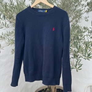Mörkblå stickad tröja Polo Ralph Lauren - Snygg mörkblå stickad tröja från Polo Ralph Lauren med klassisk röd broderad logga på bröstet. Tröjan har rund hals, ribbade muddar och långärmad passform. Perfekt för dig som gillar stilrena och tidlösa plagg.