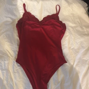 Röd spetsdetaljerad bodysuit från SHEIN - Snygg röd bodysuit från SHEIN med tunna axelband och spetsdetaljer vid bysten. Modellen är figurnära och har en djup v-ringning framtill. Perfekt att styla med jeans eller kjol för en trendig look. Passar också för xxs. Andvänts 1 gång.