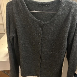 Mörkgrå kofta från 157 - Mysig mörkgrå kofta från 157 med rund halsringning och knappar framtill. Koftan är stickad och har långa ärmar, perfekt för lager på lager under kyliga dagar. Enkel och stilren design som funkar till många olika looks.