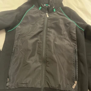 Svart vindjacka från Hugo Boss - Svart vindjacka från Hugo Boss med gröna detaljer längs axlarna och huva. Jackan har dragkedja framtill, två sidofickor med dragkedja och en snygg sportig look. Materialet är polyester och bomull, perfekt för dig som gillar stilrena och funktionella plagg.