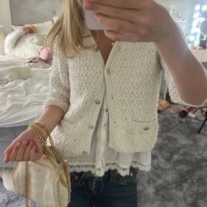 Vit populär stickad kofta med knappar - Säljer en söt vit stickad väst med dekorativa guldknappar och två små fickor fram. Västen har en grovstickad struktur och v-ringad hals. Perfekt att slänga över en blus för en chill och trendig look.