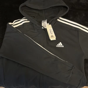 Svart Adidas hoodie med vita ränder - Svart hoodie från Adidas med klassiska vita ränder längs ärmarna och broderad logga på bröstet. Tröjan har huva, dragkedja framtill och långa ärmar. Perfekt för dig som gillar sportig stil och vill ha något bekvämt och snyggt.