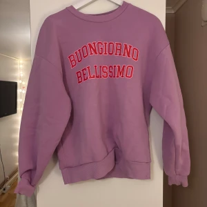 Trendig Lila sweatshirt från Gina Tricot - Cool lila sweatshirt från Gina Tricot med trycket 'BUONGIORNO BELLISSIMO' i rött och vitt på bröstet. Tröjan har ribbade muddar vid hals, ärmslut och nederkant samt en relaxed passform. Perfekt för dig som gillar färg och statement-prints.