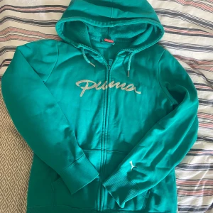 Grön hoodie från Puma med dragkedja - Turkos hoodie från Puma med broderad logga i silver på bröstet och liten Puma-symbol på ärmen. Tröjan har huva med snörning, mjukt fleecefoder och praktisk dragkedja framtill. Perfekt för chill dagar eller när du vill ha något bekvämt och snyggt.