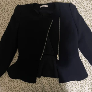 Svart kavajjacka från Mango XS - Snygg svart kavajjacka från Mango med asymmetrisk dragkedja framtill. Jackan har figursydd passform, markerad midja och långa ärmar. Perfekt för en stilren look och passar till många olika outfits.