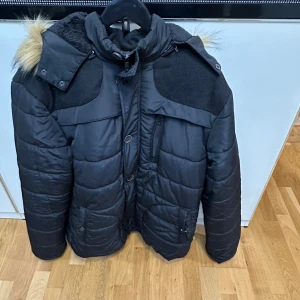 Svart pufferjacka med fuskpäls - Säljer en svart pufferjacka med mysig fuskpäls på huvan och mjukt foder på insidan. Jackan har dragkedja, knappar och flera fickor framtill. Perfekt för kalla dagar och har en riktigt skön oversized känsla. Jackan är liten på storleken och passar xl-xxl