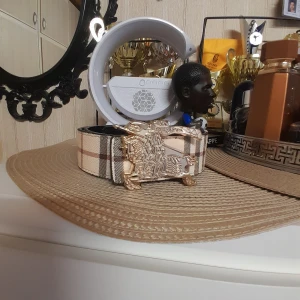 Burberry bälte - Säljer ett snyggt bälte med klassiskt rutigt mönster i beige, svart och brunt. Bältet har ett stort guldfärgat spänne med en riddare på häst. Insidan är svart och bältet är gjort i skinn. Perfekt för dig som vill ha en statement-accessoar.