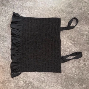 Svart smockad linnetopp från H&M - Svart smockad linnetopp från H&M med volangkant nertill och breda axelband. Toppen är ärmlös och har en tight passform som sitter snyggt på kroppen. Perfekt att matcha med jeans eller kjol för en trendig look.