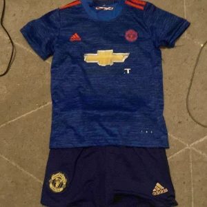 Manchester United Ibrahimovic tröja blå - Blå Manchester United fotbollströja från Adidas med röda detaljer, klubbmärke och sponsortryck fram. Nummer 9 och Ibrahimovic på ryggen. Tillhörande blå shorts med klubbmärke och Adidas-logga. Materialet är lätt och andas, perfekt för fotbollsträning.sparar till en fiddy