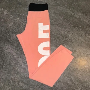 Nike rosa leggings med tryck - Nike leggings i ljusrosa med svart midja och stort vitt 'JUST DO IT'-tryck längs benet. Tight passform och stretchigt material, perfekta för träning eller chill. Snygg kontrast mellan färgerna och klassisk Nike-logga på insidan.