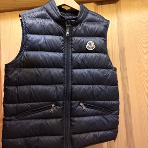 Svart dunväst från Moncler - Svart dunväst från Moncler med klassisk quiltad design, hög krage och dragkedja framtill. Västen har två snedställda fickor med dragkedja och Moncler-logga på bröstet. Yttermaterialet är i nylon och fodret är också nylon.
