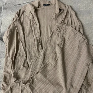 Beige dress - Beige dress  i ett mjukt, krinklat tyg med avslappnad passform. Skjortan har klassisk krage, knäppning framtill och långa ärmar. Materialet är lätt och luftigt, perfekt för varmare dagar. Säljs tillsammans med matchande byxor för en komplett look.