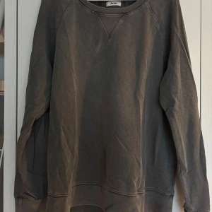 Mörkgrå sweatshirt från Acne Studios - Säljer en mörkgrå sweatshirt från Acne Studios i storlek L. Tröjan är tillverkad i 100% bomull och har klassisk rund hals med ribbade muddar och en snygg V-detalj vid halsen. Fint skick men har ett litet hål vid vänster armslut (se bild)