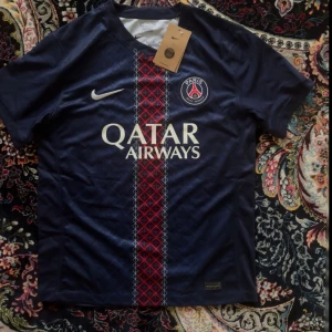 PSG matchtröja marinblå Nike - Snygg PSG fotbollströja från Nike i marinblått med röd och vit randig detalj på framsidan. Tröjan har korta ärmar, rund hals och Paris Saint-Germain-logga på bröstet. Materialet är lätt och andas, perfekt för träning eller match.