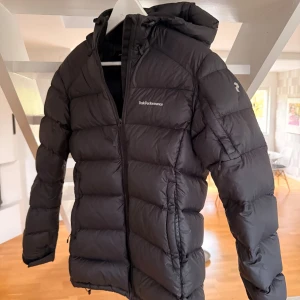 Svart dunjacka Peak Performance - Svart dunjacka från Peak Performance, modell Frost DJ. Jackan har en puffig design, huva och dragkedja framtill. Perfekt för kalla dagar och har en stilren look med diskret logga på bröstet och ärmen. Ett litet mindre hål på höger armen men syns knappt då jackan är mörk :)