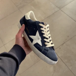 Golden Goose Superstar sneakers blå/vit - Golden Goose Superstar sneakers i blå mocka med vit stjärndetalj på sidan och vit hälkappa. Klassisk låg modell med snörning och platt sula. Tillverkade i äkta skinn och mocka, med cool vintage vibe och tydlig branding på hälen.