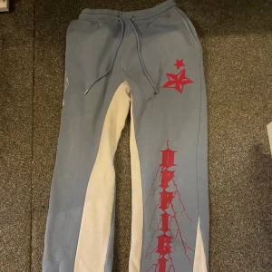 Blå mjukisbyxor med röd tryck - Snygga blå mjukisbyxor med kontrasterande beige paneler längs insidan av benen. Byxorna har snörning i midjan och coolt rött tryck med stjärna och text längs benet. Perfekt för en avslappnad streetwear-look. De är utsvängda i benen 