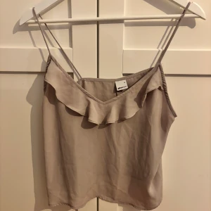 Beige linnetopp med volang från Gina Tricot - Säljer en beige linnetopp från Gina Tricot med tunna axelband och snygg volangdetalj längs ringningen. Toppen har en mjuk, flowy känsla och passar perfekt till sommaren. Enkel att matcha med både jeans och kjol.