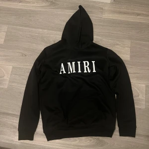 Svart hoodie Amiri - Svart hoodie Amiri med stor vit logotyp på bröstet. Tröjan har huva, lång ärm och en klassisk magficka framtill. Mjuk och skön känsla, perfekt för dig som gillar streetwear och vill ha en clean look.