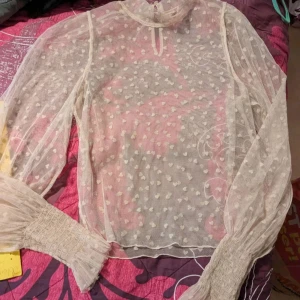 Beige meshblus med hjärtmönster - Säljer en transparent beige meshblus med små vita hjärtan över hela tyget. Blusen har långa puffärmar med smock vid handlederna och hög halsringning. Perfekt för dig som gillar romantiska detaljer och en luftig känsla. Storleken står tyvärr inte men gissar på Xs ☺️