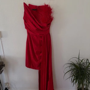 Röd asymmetrisk klänning från Day - Snygg röd långklänning från Day med en axel och fjäderdetalj upptill. Klänningen har draperad design och lång släp på ena sidan, vilket ger en dramatisk look. Perfekt för dig som vill sticka ut på bal eller fest.
