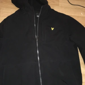 Svart vindjacka från Lyle & Scott - Svart vindjacka från Lyle & Scott med huva och gul broderad logga på bröstet. Jackan har hel dragkedja framtill och är tillverkad i ett mjukt syntetmaterial. Perfekt för dig som vill ha en stilren och enkel jacka med snygga detaljer.