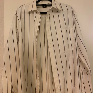 Vit randig skjorta med knappar - Vit skjorta från FSBN med svarta vertikala ränder, klassisk krage och bröstficka. Långärmad modell med knäppning framtill. Perfekt för en clean och stilren look.