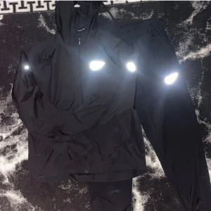 Svart Nike vindjacka med reflexdetaljer - Svart vindjacka från Nike med huva och dragkedja framtill. Jackan har tydliga reflexdetaljer och klassisk Nike-logga på bröstet. Tillverkad i lätt polyester, perfekt för blåsiga dagar. Sportig look med normal passform och långa ärmar.