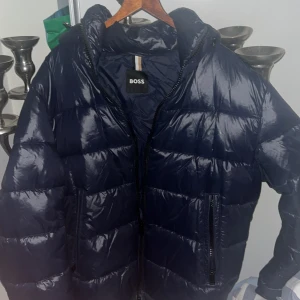 Jacka  BOSS - Säljer en mörkblå dunjacka från BOSS med glansig finish. Jackan har huva, två dragkedjefickor och quiltad design. Perfekt för kalla dagar,ny pris ca 2000kr